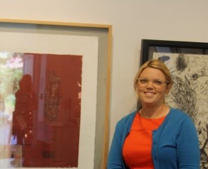 A woman stands next to her framed artwork on the wall.