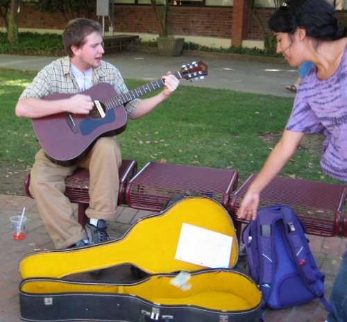 A male student plays guitar and sings as money is being tossed into his open guitar case by a woman