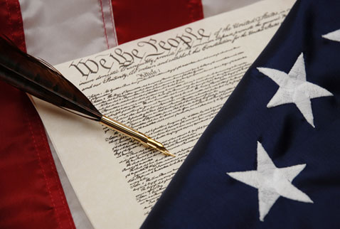 A pen and a flag lie across a printed copy of the U.S. Constitution.